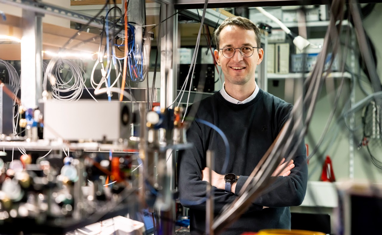 Dr. Stephan Welte steht lächelnd in einem Labor, umgeben von komplexer Laborausrüstung, Kabeln und technischen Geräten. Im Hintergrund sind weitere wissenschaftliche Instrumente und Apparaturen zu erkennen.