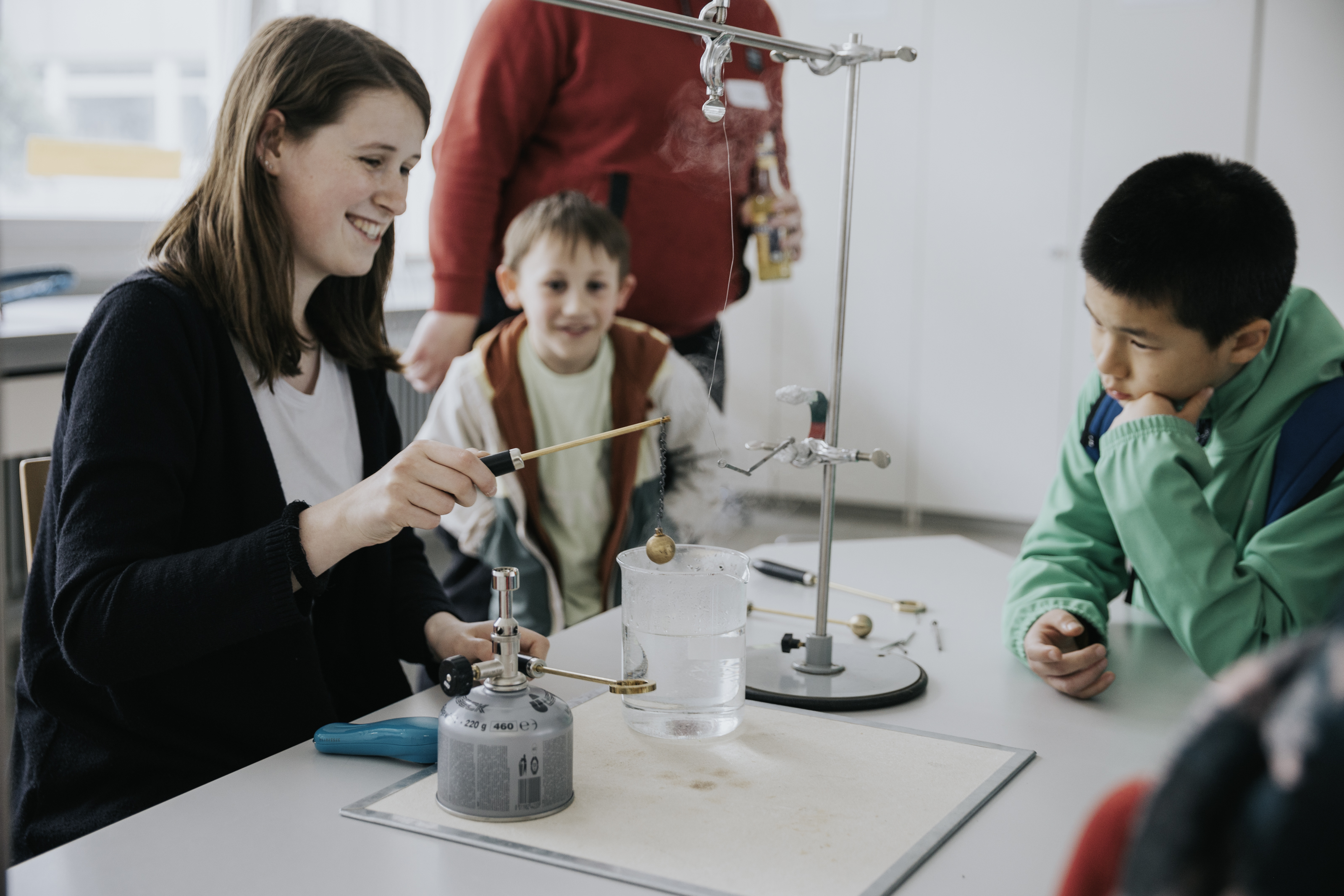 A young woman sits smiling at a table and conducts an experiment. Two young pupils watch with interest.